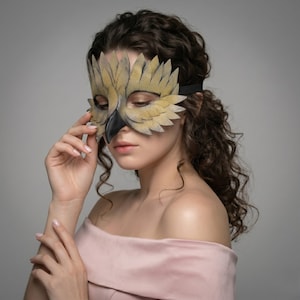 May include: A handmade owl mask in tan and black. The mask has detailed feather designs and a black beak. It is secured with a black elastic band. The mask is worn by a person wearing a pink off-the-shoulder top.