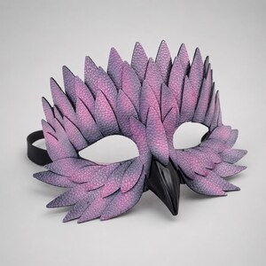 May include: A handcrafted mask with an avian theme, in shades of purple and grey, with a textured, feather-like design. The mask has a black beak and a black elastic strap. Ideal for costume parties or theatrical performances.