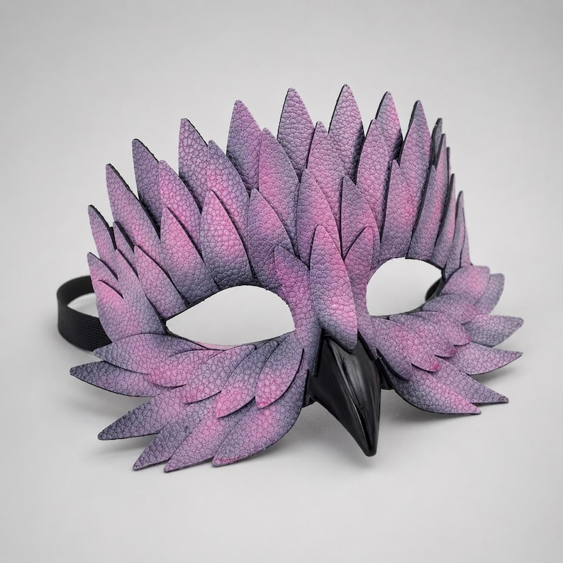 May include: A handcrafted mask with an avian theme, in shades of purple and grey, with a textured, feather-like design. The mask has a black beak and a black elastic strap. Ideal for costume parties or theatrical performances.