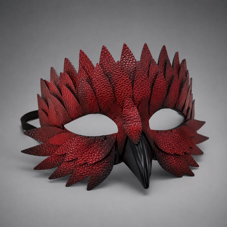 May include: A red and black masquerade mask with a textured, feathered design. The mask features a black beak and a black elastic band. The eye openings are white.