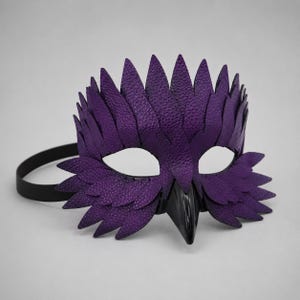 May include: A purple leather mask with a bird-like design. The mask has a textured finish, white eye openings, and a black beak. A black elastic band is attached for wear.