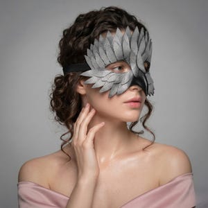 May include: A grey and black leather mask with a bird-like feather design. The mask covers the eyes and nose, with a black strap around the head. The mask is made of textured leather and has a dramatic, theatrical appearance.