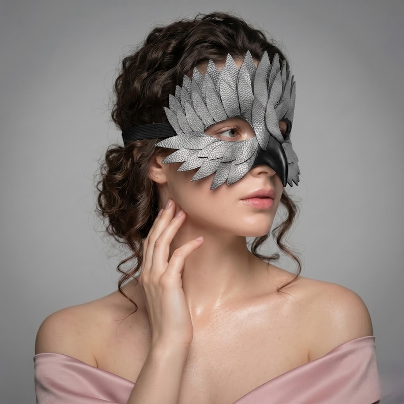 May include: A grey and black leather mask with a bird-like feather design. The mask covers the eyes and nose, with a black strap around the head. The mask is made of textured leather and has a dramatic, theatrical appearance.