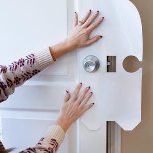 May include: A white door with a silver doorknob and a white paper template being applied. The template has cutouts for the doorknob and lock. Hands with manicured nails are touching the door and template.