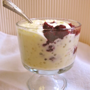 May include: A clear glass dessert dish filled with a creamy white pudding topped with a layer of dark red fruit compote. A silver spoon rests on the rim of the dish.