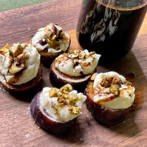 Könnte beinhalten: Nahaufnahme eines Holzbretts mit Feigenhälften, belegt mit weißem Käse, Nüssen und einem Soßenstrich. Im Hintergrund eine dunkle Glasflasche. Die Feigen sind in einer Gruppe angeordnet.
