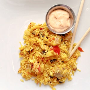 Puede incluir: Un plato blanco con una porción de arroz amarillo con verduras rojas y verdes, un pequeño tazón de salsa rosa y dos palillos de madera.