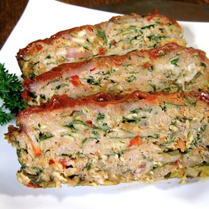 May include: A close-up of a sliced meatloaf with a red sauce on top. The meatloaf is filled with chopped vegetables, including green leafy vegetables, red peppers, and onions.