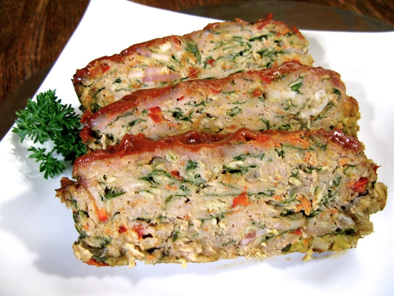 May include: A close-up of a sliced meatloaf with a red sauce on top. The meatloaf is filled with chopped vegetables, including green leafy vegetables, red peppers, and onions.