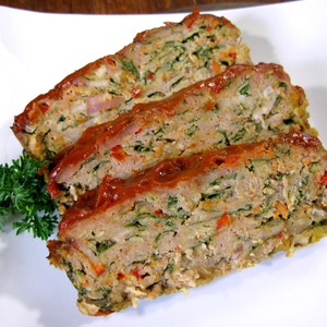 May include: Three slices of meatloaf on a white plate. The meatloaf is made with minced meat, vegetables, and herbs. It is topped with a red sauce and garnished with parsley.