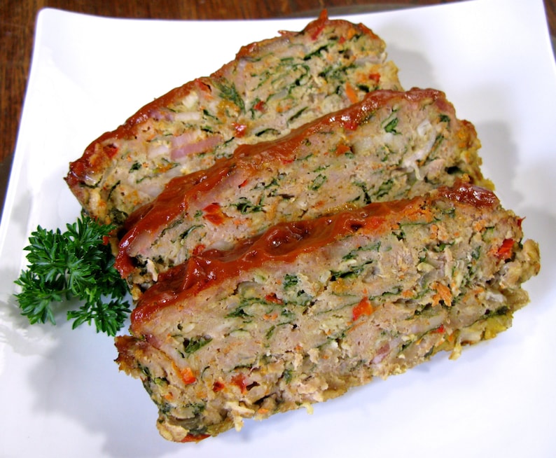 May include: Three slices of meatloaf on a white plate. The meatloaf is made with minced meat, vegetables, and herbs. It is topped with a red sauce and garnished with parsley.