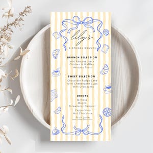 May include: A menu card with a light yellow striped background and blue decorative elements. The menu, titled "Lily's Birthday Brunch," lists brunch, sweet, and drink selections, including pancakes, waffles, and cocktails. The card is placed on a light wooden plate.