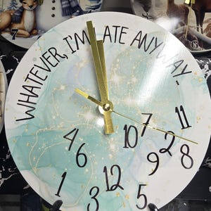 May include: A round wall clock with a white face featuring the text "WHATEVER, I'M LATE ANYWAY." The clock face has a teal and gold marbled design with black numbers and gold hands. Other clocks are visible in the background.