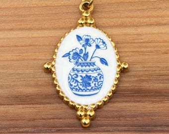 May include: A gold-toned necklace featuring an oval pendant with a white enamel center. The pendant displays a blue floral illustration of flowers in a patterned vase. The frame of the pendant has a beaded edge and is suspended from a gold chain.