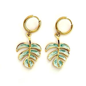 May include: A pair of gold hoop earrings featuring dangling monstera leaf charms. Each leaf charm is outlined in gold metal and filled with a marbled, swirling pattern of light green and white enamel, creating a distinct botanical appearance.