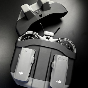 May include: A grey and black drone carrying case, open to reveal a drone and two grey battery packs. The case has a hinged lid and a moulded interior to hold the drone securely. The DJI logo is visible on the battery packs.
