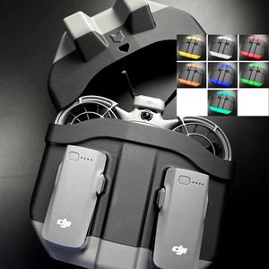 Puede incluir: Una base de carga para dron en color negro y gris con dos ranuras verticales para baterías con el logotipo de DJI y un compartimento superior. El texto MULTI-COLOUR aparece arriba, junto a una cuadrícula que muestra variantes en amarillo, blanco, rojo, naranja, azul, verde y cian.