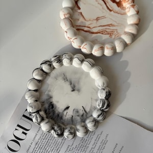 Può includere: Due vassoi decorativi con bordi perlati. Un vassoio ha un design marmorizzato bianco e marrone, e l'altro un design marmorizzato bianco e nero. I vassoi sono su una superficie bianca, con una rivista parzialmente visibile.