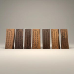 May include: Seven wooden cribbage boards in varying shades of brown. Each board is rectangular with a series of small holes for peg placement. The boards are arranged in a row, showcasing the natural wood grain and design.