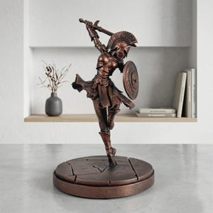 Puede incluir: Una estatua de color bronce de una figura guerrera en pose dinámica, con casco de plumas y armadura, sosteniendo una espada en alto y un escudo redondo. La figura se apoya sobre una base circular con textura de piedra agrietada.