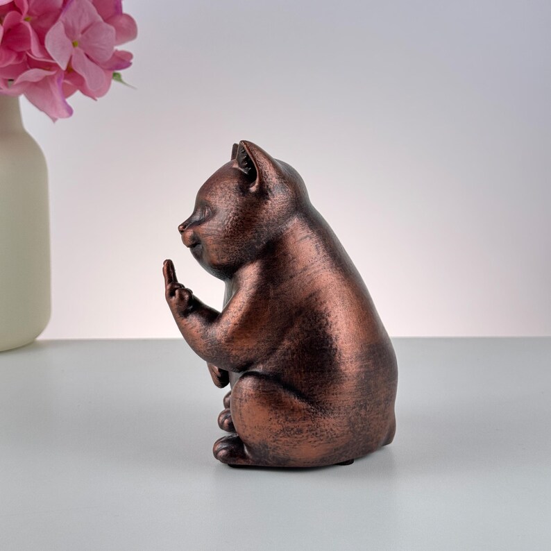 May include: A bronze-colored cat figurine in a seated position, with one paw raised, displaying the middle finger. The cat has a slightly grumpy expression. The figurine is on a white surface, with a vase of pink flowers in the background.