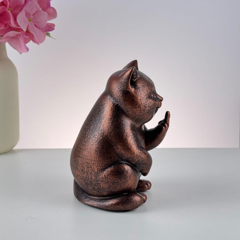 May include: A bronze-colored cat figurine in a sitting position, with one paw raised and the middle finger extended. The cat has a textured surface and is set against a neutral background. A vase with pink flowers is in the background.