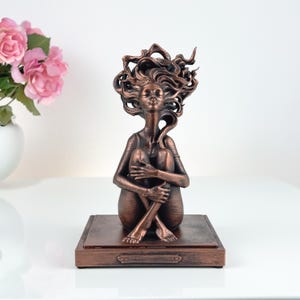 K&ouml;nnte beinhalten: Eine bronzefarbene Skulptur einer sitzenden Figur mit angezogenen Knien und geschlossenen Augen. Die Figur hat langes, flie&szlig;endes, stilisiertes Haar, das sich nach oben und au&szlig;en erstreckt. Sie ist auf einem rechteckigen Sockel mit Rand montiert.
