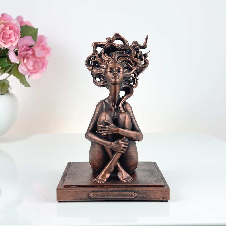 K&ouml;nnte beinhalten: Eine bronzefarbene Skulptur einer sitzenden Figur mit angezogenen Knien und geschlossenen Augen. Die Figur hat langes, flie&szlig;endes, stilisiertes Haar, das sich nach oben und au&szlig;en erstreckt. Sie ist auf einem rechteckigen Sockel mit Rand montiert.