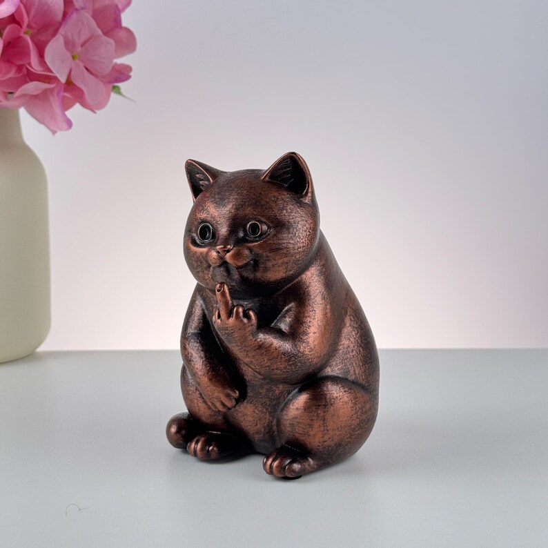 May include: Bronze-colored cat figurine with a cheeky gesture. The cat is sitting, with its paw raised, displaying the middle finger. The figurine has large, round eyes and a slightly smug expression. A humorous and unique decorative item.