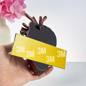 Puede incluir: Una escultura de coraz&oacute;n anat&oacute;mico de color bronce con un centro negro, sostenida en una mano. Una tira adhesiva amarilla con el texto "3M" est&aacute; adherida al coraz&oacute;n. Flores rosas en un jarr&oacute;n blanco en el fondo.