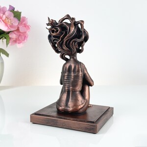 K&ouml;nnte beinhalten: Eine bronzefarbene Skulptur einer sitzenden Figur mit stilisiertem, flie&szlig;endem Haar, von hinten betrachtet. Die Figur steht auf einem rechteckigen Sockel mit demselben metallischen Finish, der eine strukturierte Oberfl&auml;che und Patina aufweist.