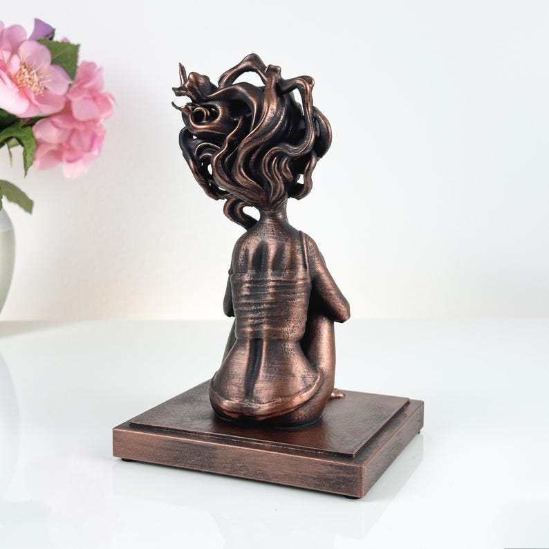 K&ouml;nnte beinhalten: Eine bronzefarbene Skulptur einer sitzenden Figur mit stilisiertem, flie&szlig;endem Haar, von hinten betrachtet. Die Figur steht auf einem rechteckigen Sockel mit demselben metallischen Finish, der eine strukturierte Oberfl&auml;che und Patina aufweist.