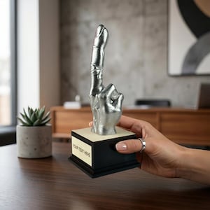 May include: A silver-colored trophy in the shape of a hand with the index finger pointing up. The trophy is mounted on a black base with a cream-colored top and the text "YOUR TEXT HERE".