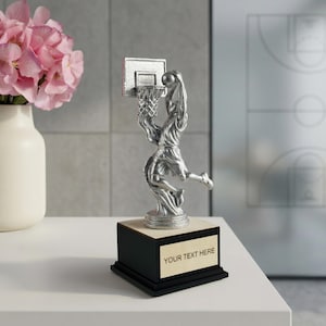 Può includere: Trofeo di basket color argento raffigurante un giocatore a metà schiacciata. Il trofeo è montato su una base nera con una placca chiara con la scritta "YOUR TEXT HERE". Un vaso di fiori rosa è sullo sfondo.