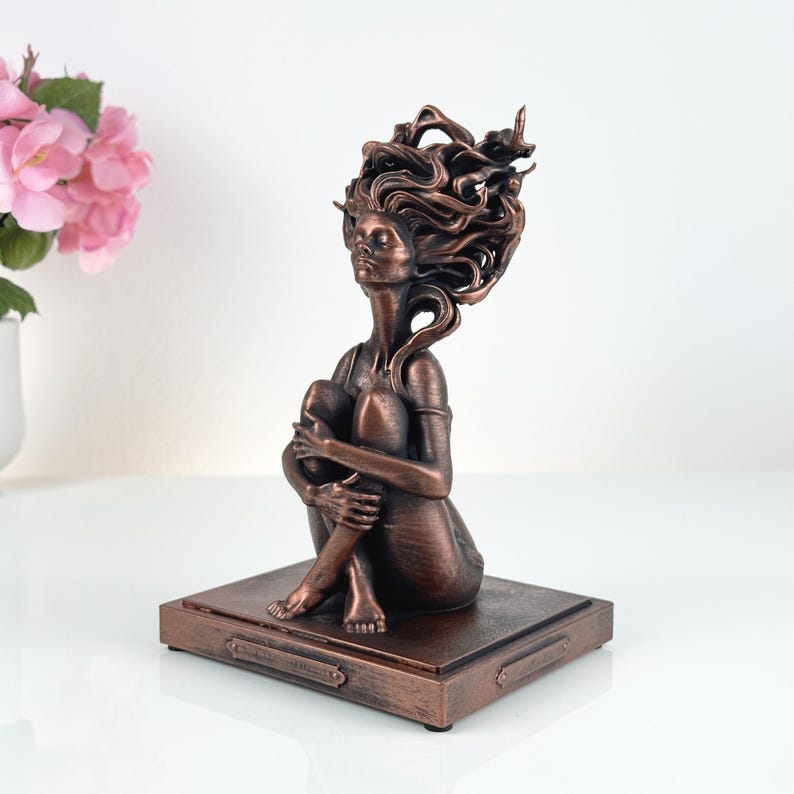 K&ouml;nnte beinhalten: Eine bronzefarbene Skulptur einer sitzenden Figur mit zum K&ouml;rper gezogenen Knien und flie&szlig;endem, stilisiertem Haar. Die Figur ist auf einem rechteckigen Sockel mit metallischem Finish montiert und f&auml;ngt eine Pose der inneren Einkehr ein.