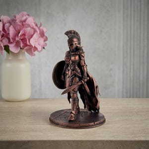 May include: A bronze-colored statue of a female warrior in full armor, holding a sword and shield. The statue has a detailed helmet and cape, standing on a round base. A vase of pink flowers is in the background.