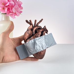 Puede incluir: Una escultura de coraz&oacute;n anat&oacute;mico de color bronce con una tira de cinta adhesiva gris. El coraz&oacute;n se sostiene en una mano, con un jarr&oacute;n de flores rosas en el fondo. La escultura tiene venas y arterias detalladas.