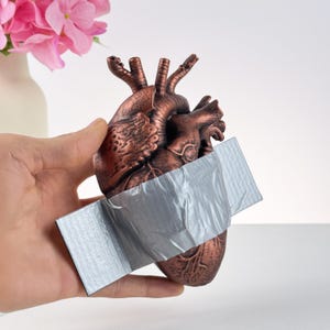 Puede incluir: Una escultura anat&oacute;mica de coraz&oacute;n de color bronce con cinta adhesiva plateada. El coraz&oacute;n est&aacute; sujeto con una mano, con un jarr&oacute;n de flores rosas en el fondo.