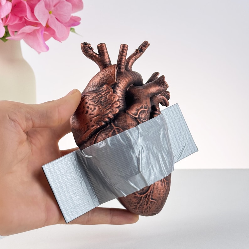 Puede incluir: Una escultura anat&oacute;mica de coraz&oacute;n de color bronce con cinta adhesiva plateada. El coraz&oacute;n est&aacute; sujeto con una mano, con un jarr&oacute;n de flores rosas en el fondo.