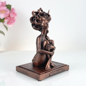 K&ouml;nnte beinhalten: Eine bronzefarbene Skulptur einer sitzenden Figur, die ihre Knie an die Brust zieht. Die Figur hat langes, flie&szlig;endes, stilisiertes Haar und ist auf einem rechteckigen Sockel montiert. Die Oberfl&auml;che hat ein strukturiertes, metallisches Aussehen.