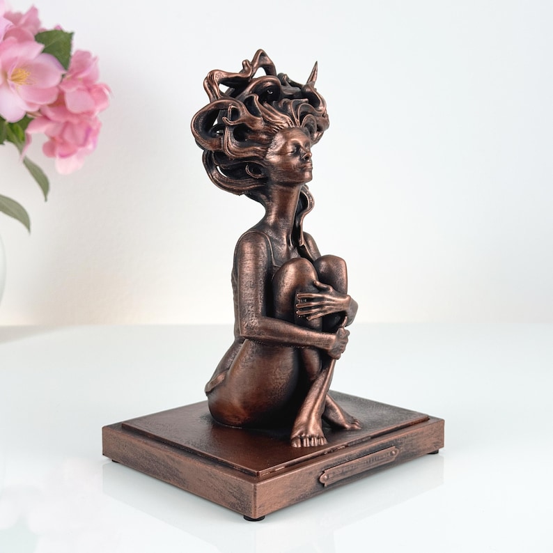 K&ouml;nnte beinhalten: Eine bronzefarbene Skulptur einer sitzenden Figur, die ihre Knie an die Brust zieht. Die Figur hat langes, flie&szlig;endes, stilisiertes Haar und ist auf einem rechteckigen Sockel montiert. Die Oberfl&auml;che hat ein strukturiertes, metallisches Aussehen.
