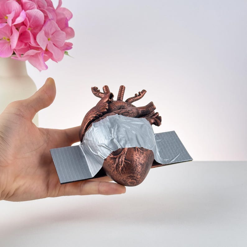 Puede incluir: Una escultura de coraz&oacute;n anat&oacute;mico de color bronce con cinta adhesiva plateada que cubre el centro. El coraz&oacute;n est&aacute; montado sobre una base gris. Flores rosas en un jarr&oacute;n blanco en el fondo.