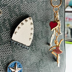 May include: A textured black surface featuring a white iron-shaped patch with silver studs, a blue circular patch with a silver star, and a gold-toned metal keychain with a red heart charm and a pink rhinestone-encrusted ballerina figure.