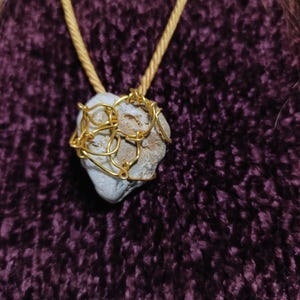 May include: A handmade necklace featuring a small, light-colored stone wrapped in gold-colored wire. The stone is suspended from a gold-colored cord, creating a simple yet elegant pendant. The background is a dark purple, textured fabric.