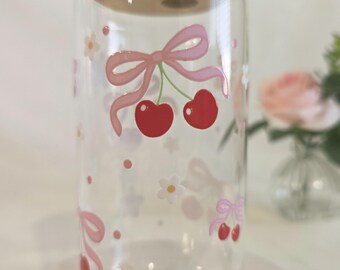 May include: A clear glass tumbler with a bamboo lid featuring a repeating pattern of red cherries tied with light pink bows, interspersed with small white flowers and pink dots. The design is printed directly onto the exterior of the glass.