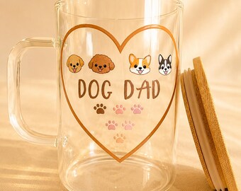 May include: A clear glass mug with a handle and a bamboo lid. The front features a brown heart outline containing illustrations of four dog faces, the text "DOG DAD" in brown letters, and a series of small brown and pink paw print icons below.