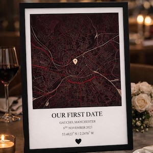 May include: A framed map print with the text "OUR FIRST DATE" and location details. The map is in burgundy and gold tones, with a pin marking a specific spot. The frame is black, and the print includes the date and coordinates.