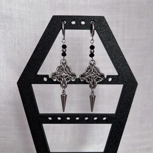 May include: A pair of silver-toned earrings displayed on a black earring stand. Each earring features a chainmail-style design with a dangling spike and a strand of black beads. The earrings are suspended from hoop closures.