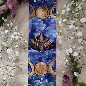 May include: A rectangular bookmark with a blue and purple background, featuring a moth illustration and two gold crescent moon designs. The bookmark is surrounded by white and purple flowers.