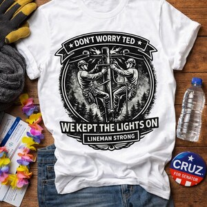 Op de afbeelding: Wit T-shirt met een afbeelding van twee elektriciens op een elektriciteitspaal, met de tekst "DON'T WORRY TED, WE KEPT THE LIGHTS ON, LINEMAN STRONG." Het shirt ligt op een houten oppervlak met andere items.